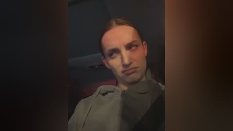 Media: Video of a Caucasian man with short brown hair, wearing a gray jacket, seated in a dimly lit car interior, with blurred surroundings.