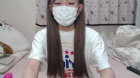 Media: Video of an Asian woman with long brown hair wearing a white T-shirt, blue face mask, and small earrings, seated in a messy bedroom with white sheets and a floral-patterned curtain.