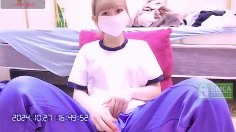 Media: Video of a young Asian girl with light skin, wearing a white T-shirt, blue sweatpants, and a face mask, seated on a bed with a pink pillow, in a tidy bedroom.