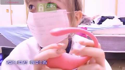 Media: Video of a young woman wearing a pink face mask, holding a pink vibrator, in a bedroom with a bed and clothes visible.