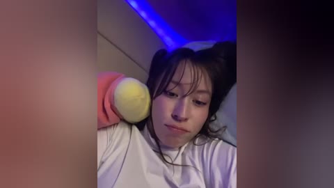 Media: Video of a young Asian woman with dark hair in buns, wearing a white turtleneck, holding a yellow tennis ball, set against a dimly lit room with blue LED lights.