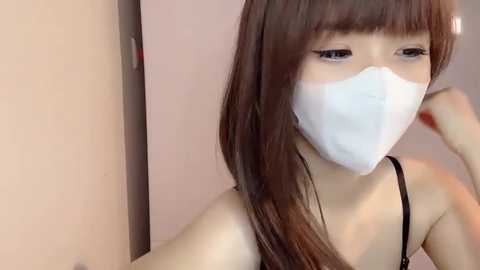 Media: A video of an Asian woman with long brown hair, wearing a white surgical mask, black tank top, and makeup. She has fair skin and is indoors with a beige wall and a door in the background.