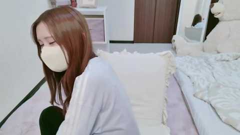 Media: Video of a young Asian woman with long brown hair, wearing a white mask, sitting on a bed with white bedding, in a modern, minimally decorated bedroom.