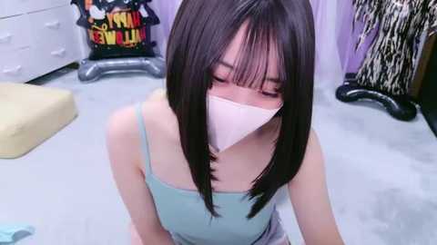 Media: A video of an Asian woman with straight black hair, wearing a light blue spaghetti-strap top, a white face mask, and sitting on a carpeted floor in a room with purple curtains, a white dresser, and a chair.