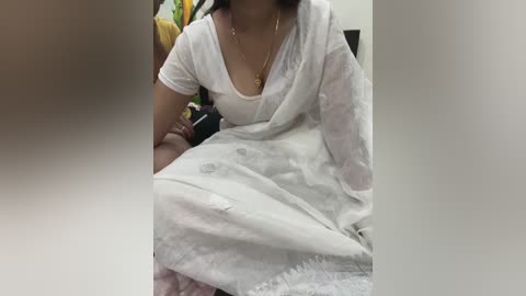 Media: Video of a woman in a white sari with a V-neck blouse, sitting with her arms crossed, wearing a gold necklace. Background shows blurred household objects.