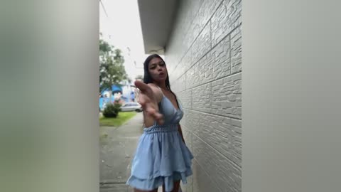 Media: Video of a young Asian woman in a blue sundress, standing against a white brick wall, extending her hand toward the camera, surrounded by a narrow corridor.