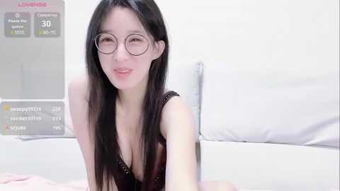 Media: A video of a young Asian woman with long black hair, wearing glasses and a black lace bra, leaning forward in a white room with a bed.