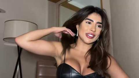 Media: Video of a Latina woman with long dark hair, fair skin, wearing a black leather bralette, adjusting her hair in a modern, minimalist room with a beige wall and floor lamp.