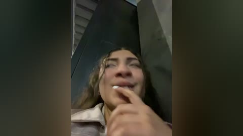 Media: Video of a young woman with long, wavy brown hair, light skin, and a white shirt, smiling and biting her fingernail, taken from a low angle.