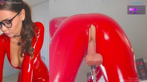 Media: Video of a woman with long brown hair, wearing black-rimmed glasses and a tight red latex bodysuit, bending over, exposing her buttocks and vagina with a large dildo inserted.