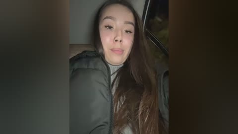 Media: Video of a young Asian woman with long brown hair, wearing a dark jacket, sitting in a car, making a duck face.