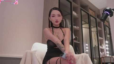 Media: Video of a tattooed, fair-skinned woman with long black hair in a black, sheer dress, sitting in a dressing room with mirrored wardrobes and a white chair.