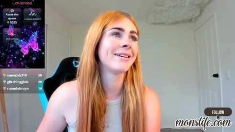 Media: Video of a smiling, fair-skinned young woman with long, straight blonde hair, wearing a white tank top, seated in a black gaming chair. Background shows a room with white walls and a closed door.