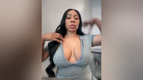 Media: Video of a plus-sized Black woman with long black hair, wearing a low-cut, light blue top that accentuates her large breasts. She stands in a modern bathroom with white tiles, holding her hair with one hand.