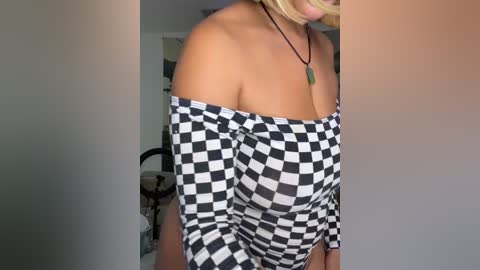 Media: A video of a light-skinned woman with blonde hair, wearing a black and white checkered off-shoulder dress, standing in a dimly lit room with blurred background details.