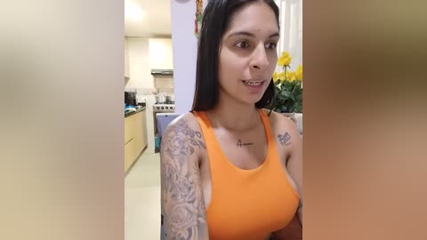 Media: Video of a Latina woman with medium skin tone and long black hair, wearing an orange tank top. She has intricate tattoos on her left arm and chest. The background shows a modern kitchen with light walls, cabinets, and a flower arrangement.