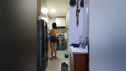 Media: Video of a petite, dark-skinned woman with long hair, wearing an orange sports bra and shorts, cooking in a small, dimly lit kitchen with a stainless steel stove, a fridge, and cluttered countertops.