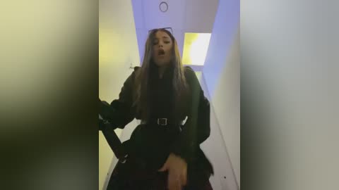 Media: Video of a young woman with long, straight brown hair, wearing a dark coat and black skirt, standing in a dimly lit, narrow hallway. She appears distressed, with her mouth open and hand to her cheek.