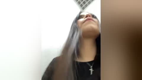 Media: Video of a young woman with long dark hair, wearing a black top and a silver cross necklace, looking up at a ceiling with visible grid pattern, giving a sense of upward motion.