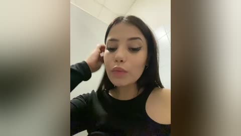 Media: Video of a young Latina woman with long black hair and fair skin, wearing black clothing, applying makeup in a bathroom with white walls and a ceiling light.