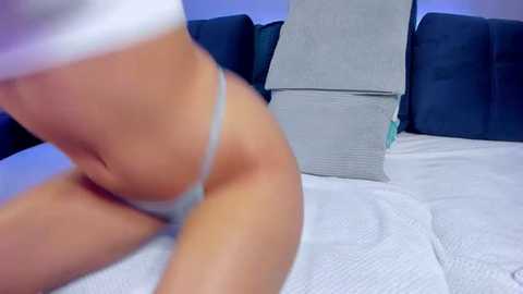 Media: Video of a person wearing light grey thong underwear, sitting on a white bed with dark blue pillows and grey box in the background.