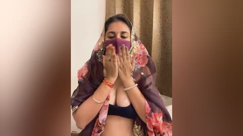 Media: Video of a woman wearing a maroon floral saree, covering her face with her hands, revealing a black bra underneath. She has a medium complexion and wears a red bangle on her right wrist.