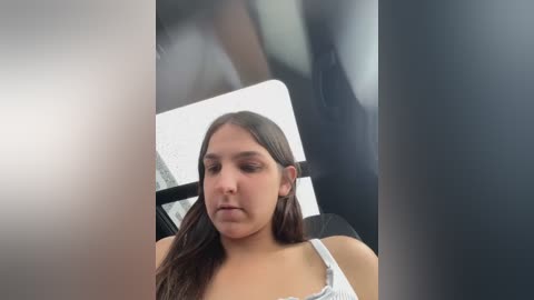 Media: A video of a young Latina woman with long, straight brown hair, wearing a white spaghetti-strap top, sitting in a car with blurred background.