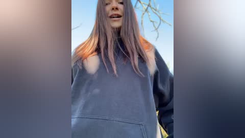 Media: Video of a young woman with long, straight, light brown hair, wearing a large, baggy black hoodie, partially visible breasts, standing outdoors with a clear blue sky and leafless tree branches in the background.