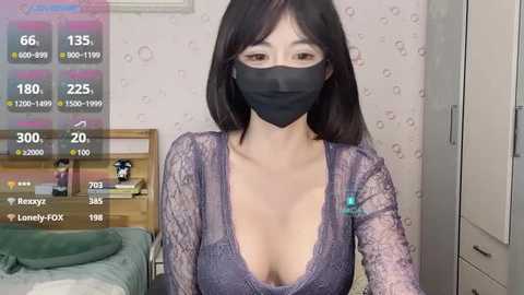 Media: A video of a young Asian woman with long black hair, wearing a black face mask and a low-cut, sheer purple lace top, indoors, with a green bed and white wallpaper in the background.