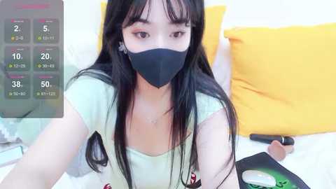 Media: A video of an East Asian woman with long black hair, wearing a black face mask and green top, lying on a bed with yellow pillows, holding a smartphone.