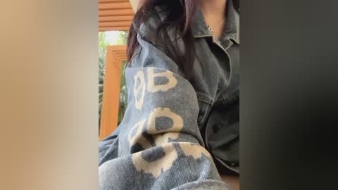 Media: Video of a woman with long, dark hair, wearing a gray denim jacket over a black shirt, sitting on a wooden chair with a blue, floral-patterned blanket draped over her lap. The background features a wooden door and greenery outside.