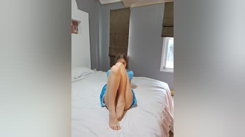 Media: A video of a young woman with light skin and brown hair, nude from the waist down, sitting on a bed with white sheets, wearing a blue top, in a gray-painted room with a window.