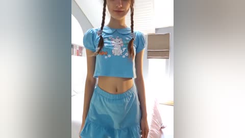 Media: Video of a slender, fair-skinned woman with long, braided brown hair, wearing a blue crop top and matching high-waisted skirt with a cartoon character print. She stands in a brightly lit, minimalist bedroom with white walls and a bed in the background.