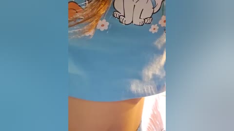 Media: A video showing a close-up of a young girl's torso wearing a blue T-shirt with a cartoon cat and white flowers. The background is a clear blue sky.