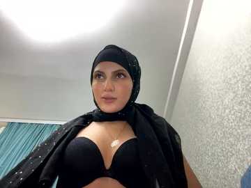 Media: Video of a light-skinned woman with medium-length hair, wearing a black hijab and matching bra, standing indoors against a textured white wall and teal curtain.