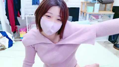 Media: Video of a young Asian woman with shoulder-length brown hair, wearing a pink ribbed sweater and a white face mask, taking a selfie in a cluttered room with clothes hanging and a desk in the background.