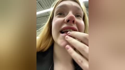 Media: Video of a young, fair-skinned, blonde woman with a nose ring, smiling and licking her finger, in a dimly lit room with metallic ceiling panels.