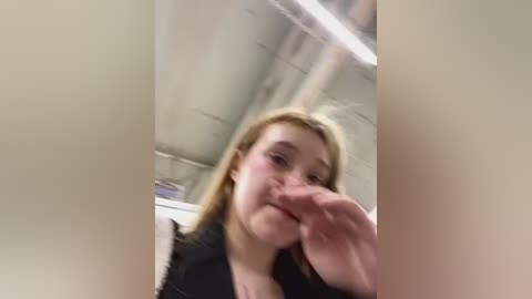 Media: Video of a young woman with light skin, straight blonde hair, and a slim build, wearing a black top, licking her fingers in a casual indoor setting with exposed concrete ceiling.