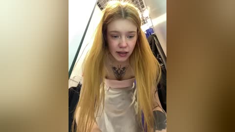 Media: A video of a young Caucasian woman with long, straight blonde hair and a pink top, leaning forward, with a tattoo on her chest, in a cluttered, beige-toned room with hanging clothes and a ceiling light.