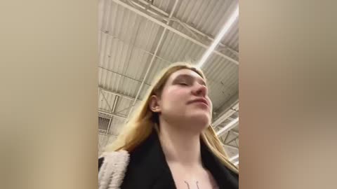 Media: Video of a young woman with long, straight blonde hair, wearing a black jacket, looking upwards with a serene expression. She stands in a large, industrial room with a high, ribbed ceiling and fluorescent lights.