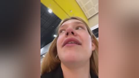 Media: A video of a smiling young woman with light skin and long, straight brown hair, captured from a low angle. She has a slightly flushed complexion. The background features a yellow ceiling and a blurred, dark-skinned person on the left.