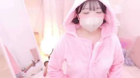 Media: A young Asian woman with fair skin, wearing a fluffy pink hoodie and white face mask, stands in a softly lit room with pastel decor.