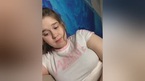 Media: A video of a young woman with light skin, wearing a white t-shirt with pink text, lying on a bed with a blue and beige backdrop.