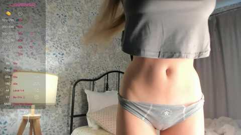 Media: Video of a young woman in gray crop top and panties, standing in a dimly lit bedroom with floral wallpaper and a metal-framed bed.