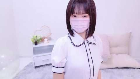 Media: Video of an East Asian woman with short, dark hair and a white surgical mask, wearing a white blouse with black trim, standing in a minimalist, bright room with a white couch, a white nightstand, and a small plant.