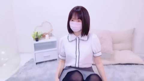 Media: Video of an Asian woman with short black hair, wearing a white blouse, gray skirt, and black stockings, sitting on a bed with white pillows, in a bright, minimalist room with white walls.