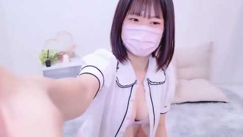 Media: A video of an Asian woman with straight black hair, wearing a white surgical mask, taking a selfie in a minimalistic, modern bedroom. She's partially dressed in a white blouse, exposing cleavage.