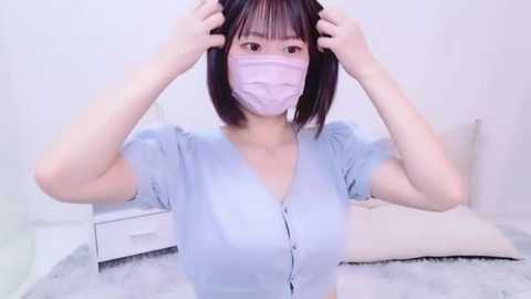 Media: Video of an East Asian woman with shoulder-length black hair, wearing a light blue button-down shirt, a pink face mask, and adjusting her headband. She's on a fluffy white rug in a minimalist, light-colored room.