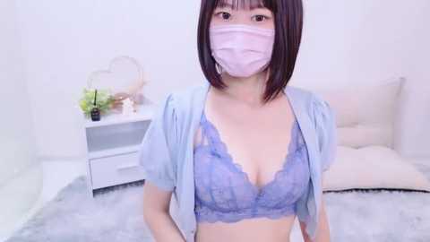 Media: Video of an East Asian woman with straight dark brown hair and a light complexion, wearing a pink mask, light blue shirt, and a lacy lavender bra, standing in a minimalist, white-walled bedroom with a fluffy gray rug.