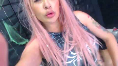 Media: Video of a fair-skinned woman with long, wavy pink hair, wearing a black Spider-Man costume with white webbing, sitting on a dark bed. She has a slightly pouty expression and visible tattoos on her arms.